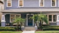 A violet craftsman-style house in Tampa with a dark blue, paned-glass front door, and small palm trees framing the entrance to the house.